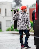 A person is wearing a patterned jacket, jeans, knee pads, and a red helmet while carrying a skateboard over their shoulder. They stand on a wet pavement with blurred buildings and trees in the background.
