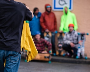 A group of people wearing colorful clothing stand close to each other against a light-colored wall. One person in the foreground holds a yellow jacket. There are skateboards visible near them, and a disability parking sign is mounted on the wall.