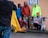 A group of people wearing colorful clothing stand close to each other against a light-colored wall. One person in the foreground holds a yellow jacket. There are skateboards visible near them, and a disability parking sign is mounted on the wall.