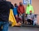 A group of people wearing colorful clothing stand close to each other against a light-colored wall. One person in the foreground holds a yellow jacket. There are skateboards visible near them, and a disability parking sign is mounted on the wall.