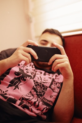 A person is relaxing on a red couch, focused on holding and looking at a handheld gaming device. They are wearing a black t-shirt with a vibrant pink and black graphic design.