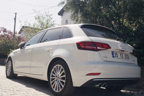 A white Audi A3 is parked on a paved driveway surrounded by greenery and a white house. The car's design features include sleek tinted windows, alloy wheels, and dual exhaust pipes. The license plate indicates a Turkish registration. In the background, there are trees and flowering shrubs adding a serene and residential atmosphere.