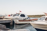 Two motorboats docked at a wooden pier in a calm harbor. One of the boats has the name 'HAYALIM' written on its side. Both boats are white with blue detailing and have Turkish flags on them. The backdrop includes a serene body of water, small hills, and a clear sky.