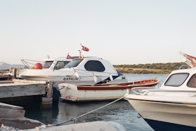 Two motorboats docked at a wooden pier in a calm harbor. One of the boats has the name 'HAYALIM' written on its side. Both boats are white with blue detailing and have Turkish flags on them. The backdrop includes a serene body of water, small hills, and a clear sky.