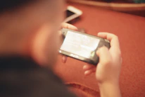 A person is holding a gaming device while seated on a reddish surface. A smartphone is placed on the table in the background. The image is out of focus, giving a soft, blurred effect.