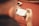 A person is holding a gaming device while seated on a reddish surface. A smartphone is placed on the table in the background. The image is out of focus, giving a soft, blurred effect.