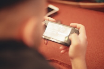 A person is holding a gaming device while seated on a reddish surface. A smartphone is placed on the table in the background. The image is out of focus, giving a soft, blurred effect.