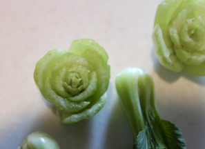 Green vegetables arranged on a light surface, including a carved piece resembling a flower. The texture and shape of the vegetables suggest freshness.