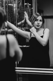 A woman in an elegant dress stands in front of a mirror, with one hand touching her neck and the other arm resting on the mirror. The setting appears introspective and intimate, with a focus on her expression and the play of reflections.