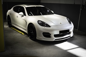 A sleek white sports car is parked in an indoor garage with soft lighting highlighting its curves and glossy finish. The car features black rims and a low profile grille, indicative of high performance vehicles. The setting is minimalistic and dimly lit, contributing to a luxurious and elegant atmosphere.