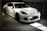 A sleek white sports car is parked in an indoor garage with soft lighting highlighting its curves and glossy finish. The car features black rims and a low profile grille, indicative of high performance vehicles. The setting is minimalistic and dimly lit, contributing to a luxurious and elegant atmosphere.