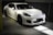 A sleek white sports car is parked in an indoor garage with soft lighting highlighting its curves and glossy finish. The car features black rims and a low profile grille, indicative of high performance vehicles. The setting is minimalistic and dimly lit, contributing to a luxurious and elegant atmosphere.