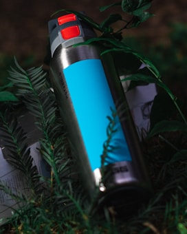 A stainless steel insulated bottle with a blue label and a red flip-top lid is situated among green foliage. The bottle appears to be designed for keeping liquids hot or cold and is partially obscured by leaves, suggesting an outdoor setting.