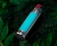 A metal water bottle with a turquoise stripe, featuring a red button on the cap, lies amidst lush, dark green foliage.