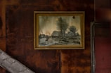 A framed painting is mounted on a rust-colored wall, depicting a pastoral scene with trees, a river, and traditional thatched-roof buildings. The wall has a weathered and industrial appearance with diagonal wooden beams.