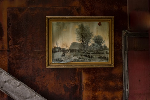 A framed painting is mounted on a rust-colored wall, depicting a pastoral scene with trees, a river, and traditional thatched-roof buildings. The wall has a weathered and industrial appearance with diagonal wooden beams.
