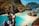 A couple stands atop a rocky viewpoint overlooking a stunning tropical bay. The scene is dominated by clear turquoise waters and rugged limestone cliffs, covered in lush green vegetation with touches of orange. The man is wearing a cap and shorts, while the woman is in a bikini, capturing the essence of a perfect vacation moment.