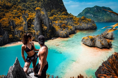 A couple stands atop a rocky viewpoint overlooking a stunning tropical bay. The scene is dominated by clear turquoise waters and rugged limestone cliffs, covered in lush green vegetation with touches of orange. The man is wearing a cap and shorts, while the woman is in a bikini, capturing the essence of a perfect vacation moment.