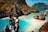 A couple stands atop a rocky viewpoint overlooking a stunning tropical bay. The scene is dominated by clear turquoise waters and rugged limestone cliffs, covered in lush green vegetation with touches of orange. The man is wearing a cap and shorts, while the woman is in a bikini, capturing the essence of a perfect vacation moment.