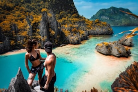 A couple stands atop a rocky viewpoint overlooking a stunning tropical bay. The scene is dominated by clear turquoise waters and rugged limestone cliffs, covered in lush green vegetation with touches of orange. The man is wearing a cap and shorts, while the woman is in a bikini, capturing the essence of a perfect vacation moment.