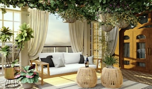 A cozy, elegantly-decorated sunroom or patio area with a natural and earthy vibe. It features a wooden-framed couch with white cushions, flanked by tall, lush green potted plants. Above, hanging plants with greenery and flowers create a canopy effect. The space is enclosed with glass doors and light-filtering curtains, letting in soft natural light. Two round wicker tables hold a bottle and glasses, adding a touch of sophistication.
