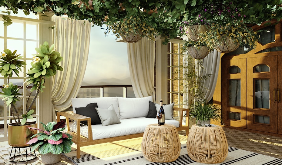 A cozy, elegantly-decorated sunroom or patio area with a natural and earthy vibe. It features a wooden-framed couch with white cushions, flanked by tall, lush green potted plants. Above, hanging plants with greenery and flowers create a canopy effect. The space is enclosed with glass doors and light-filtering curtains, letting in soft natural light. Two round wicker tables hold a bottle and glasses, adding a touch of sophistication.