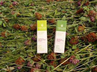Two rectangular packages of Lamacoppa Leaf Sciences full spectrum hemp extract lie on a bed of green foliage and red clover flowers. The packaging is primarily green and white with floral designs and text indicating the product details.