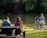 A family sits and stands near a serene lake surrounded by dense greenery. The water reflects the trees, and sunlight casts gentle shadows. Two children stand close to the water's edge while the adults sit on a wooden bench.