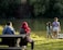 A family sits and stands near a serene lake surrounded by dense greenery. The water reflects the trees, and sunlight casts gentle shadows. Two children stand close to the water's edge while the adults sit on a wooden bench.