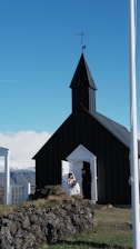 A small black church with a pointed roof stands against a clear blue sky. A bride and groom are stepping out of the doorway, with the bride in a white dress holding a bouquet of flowers. The foreground features a rocky area with moss and grass. Snow-capped mountains are visible in the background.