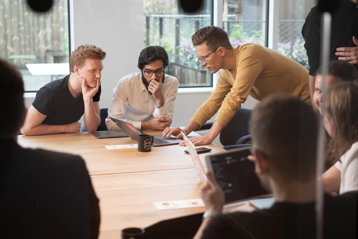 A man in a mustard sweater leans over a meeting table to walk two seated colleagues through something on an open laptop