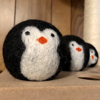 Three felted wool balls are crafted to resemble penguins, featuring distinctive black, white, and orange colors. They are lined up on a wooden surface, each displaying a black back, white face, and orange beak.