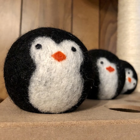 Three felted wool balls are crafted to resemble penguins, featuring distinctive black, white, and orange colors. They are lined up on a wooden surface, each displaying a black back, white face, and orange beak.