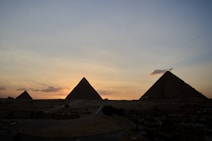 Silhouettes of several pyramids are set against a colorful sunset sky. The image captures a serene and majestic view of the pyramids, with the sun setting in the background, casting a warm glow over the sky and clouds. The ground is mostly in shadow, creating a juxtaposition between the light and dark elements.