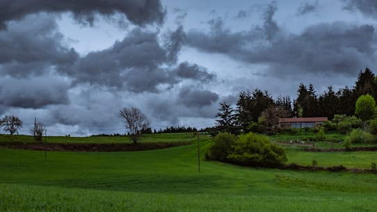 A lush, green landscape with rolling hills and a backdrop of dark, ominous storm clouds. A small house is nestled among trees on the right side, with fields and trees scattered across the scene. Power lines cut across the landscape, and the overall atmosphere is one of impending rain.