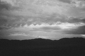 A black and white photograph features a rugged mountain landscape silhouetted against a sky filled with textured clouds. The image appears grainy, conveying a nostalgic or classic feel.