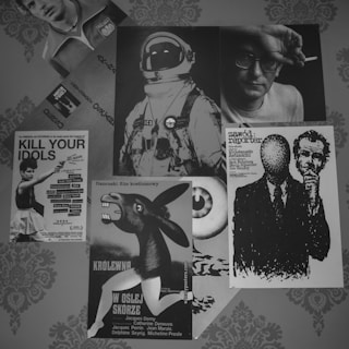 Multiple posters are displayed on a patterned textured wall. In the center, an image of an astronaut in classic gear is prominent. To the right, there is a black-and-white illustration of two men, one with an indistinct, texture-covered face. Below this, there's a poster with text in another language featuring a surreal image of a person with a donkey's head. In the top left, a face of a person can be seen, while directly below it, a monochrome photograph shows a person's face wearing glasses and holding a cigarette. There are two other posters visible, one with a dancer and bold lettering 'Kill Your Idols' and an image resembling an eyeball.