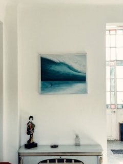 A room corner featuring a minimalist decor. On the wall, there is a painting of a large ocean wave in blue tones. Below the painting, a small white console table holds a decorative figurine of a person in traditional attire, a round dish, and a clear bottle. A section of a door with glass panels is visible to the right.
