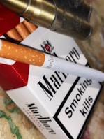 A pack of Marlboro cigarettes is prominently displayed, accompanied by a single cigarette lying on top. A warning label that reads 'Smoking kills' is visible on the package. Metallic bullet casings are seen adjacent to the cigarettes. The background is a textured surface with muted colors.