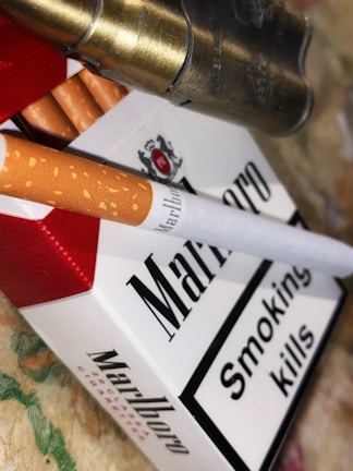 A pack of Marlboro cigarettes is prominently displayed, accompanied by a single cigarette lying on top. A warning label that reads 'Smoking kills' is visible on the package. Metallic bullet casings are seen adjacent to the cigarettes. The background is a textured surface with muted colors.