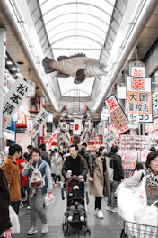 A bustling market scene with numerous people walking around. Suspended from the ceiling is a large fish, and a variety of signs with Japanese characters are hung, promoting different stores and products. The market is enclosed with a high, arched glass roof.