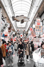 A bustling market scene with numerous people walking around. Suspended from the ceiling is a large fish, and a variety of signs with Japanese characters are hung, promoting different stores and products. The market is enclosed with a high, arched glass roof.