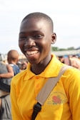 A smiling person wearing a bright yellow polo shirt with 'Wechau Girl's Conference' written on it is standing outdoors. Other people are visible in the background, suggesting a public gathering or event.