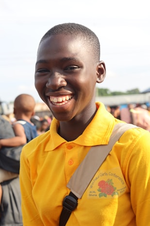 A smiling person wearing a bright yellow polo shirt with 'Wechau Girl's Conference' written on it is standing outdoors. Other people are visible in the background, suggesting a public gathering or event.