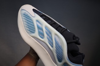 A sneaker with a futuristic design featuring a textured blue and white upper, layered on a cream sole. The shoe has a sleek, modern aesthetic with intricate details and various materials visible.