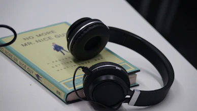 A pair of black headphones rests on top of a paperback book with the title 'No More Mr. Nice Guy' by Glover. The book is positioned on a white surface, and its cover showcases a minimalist design with text and an illustration in the center.