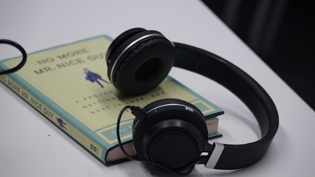 A pair of black headphones rests on top of a paperback book with the title 'No More Mr. Nice Guy' by Glover. The book is positioned on a white surface, and its cover showcases a minimalist design with text and an illustration in the center.