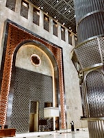 A grand architectural interior showcasing intricate geometric patterns and ornate designs. Large vertical panels with a lattice structure in metallic tones adorn the walls, complemented by a central decorative element featuring a circular motif. The area is spacious and features a high ceiling with numerous lights.
