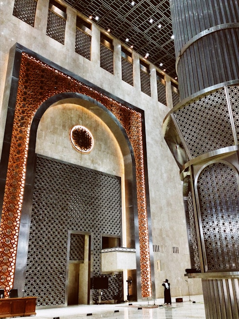 A grand architectural interior showcasing intricate geometric patterns and ornate designs. Large vertical panels with a lattice structure in metallic tones adorn the walls, complemented by a central decorative element featuring a circular motif. The area is spacious and features a high ceiling with numerous lights.
