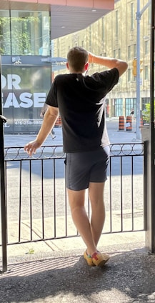 A person stands on a terrace, gazing out through a metal railing at a city street. They are wearing a black t-shirt, dark shorts, and colorful shoes. A large building with glass windows and an 'For Lease' sign is visible in the background, along with orange traffic cones.
